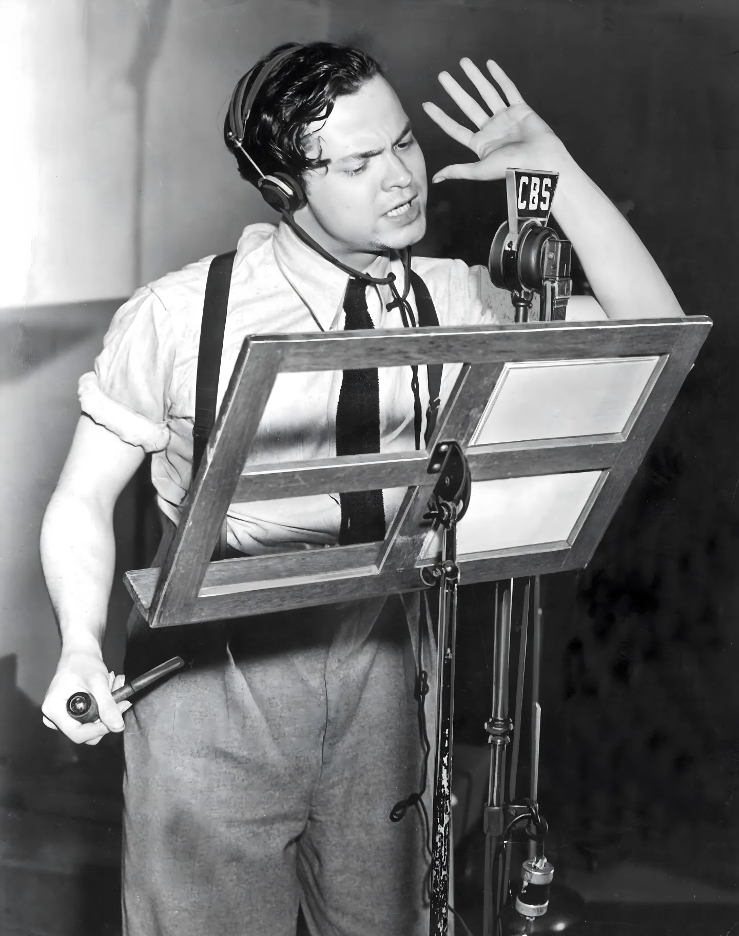 Orson Welles directing in a CBS radio studio, 1938; leaning into a ribbon microphone with one hand raised, mid-cue.